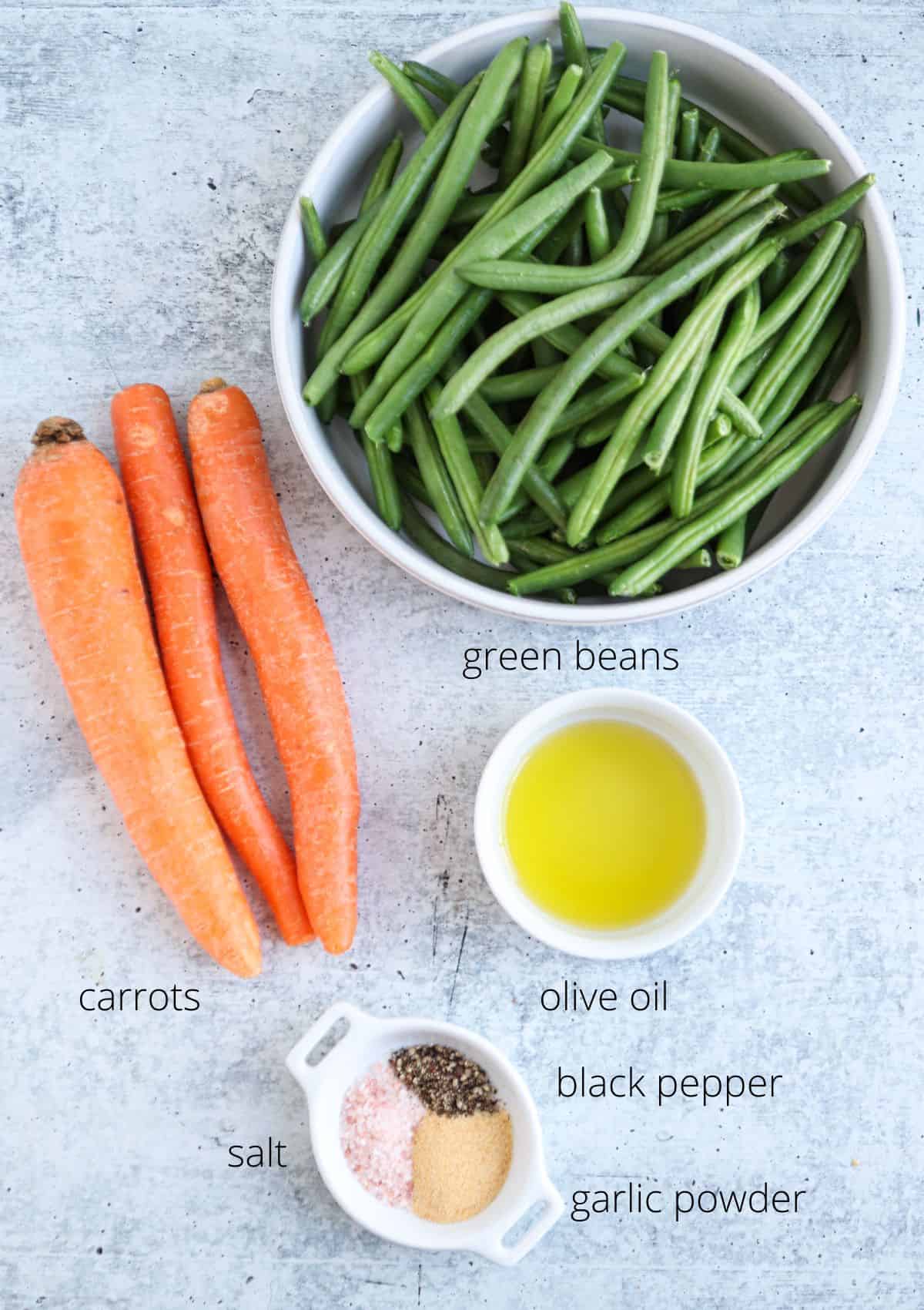 A top view of fresh green beans in a bowl, three carrots, and a small bowl of olive oil, complete with seasonings like black pepper, salt, and garlic powder on a light textured background. Each item is labeled for easy identification.