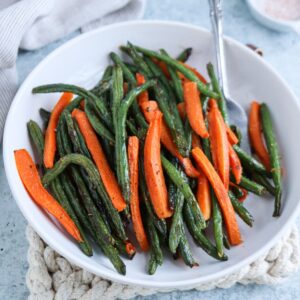 A white plate filled with roasted green beans and carrot sticks, seasoned with herbs and pepper. A fork rests on the plate, while a woven mat lies underneath. A cloth and a small dish add charm in the background.