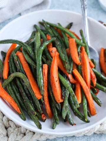 A white plate filled with roasted green beans and carrot sticks, seasoned with herbs and pepper. A fork rests on the plate, while a woven mat lies underneath. A cloth and a small dish add charm in the background.