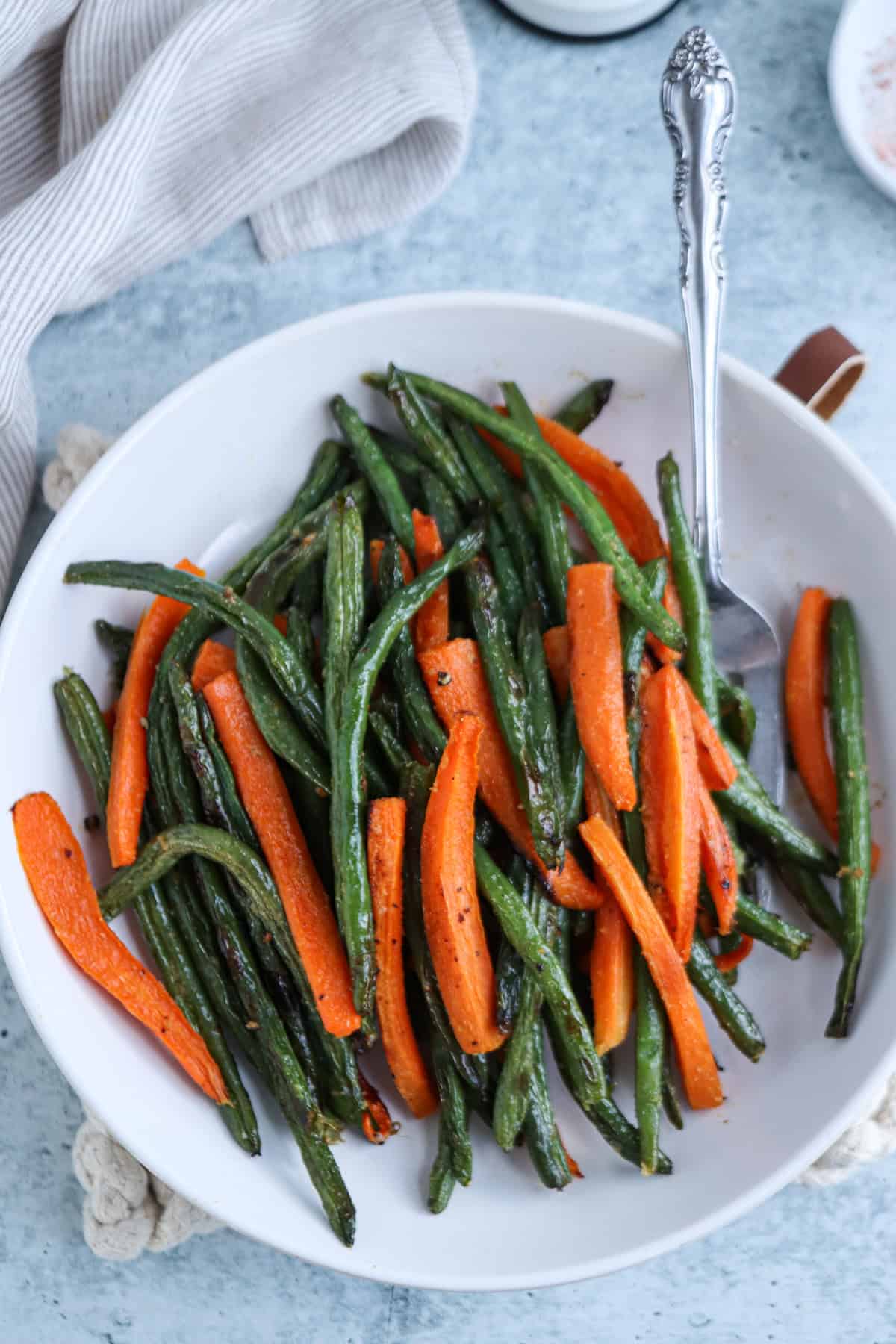 A white dish filled with roasted green beans and carrot sticks, is lightly garnished. A silver spoon rests in the dish. The background is a textured blue-gray surface with a glimpse of a light gray cloth in the corner.