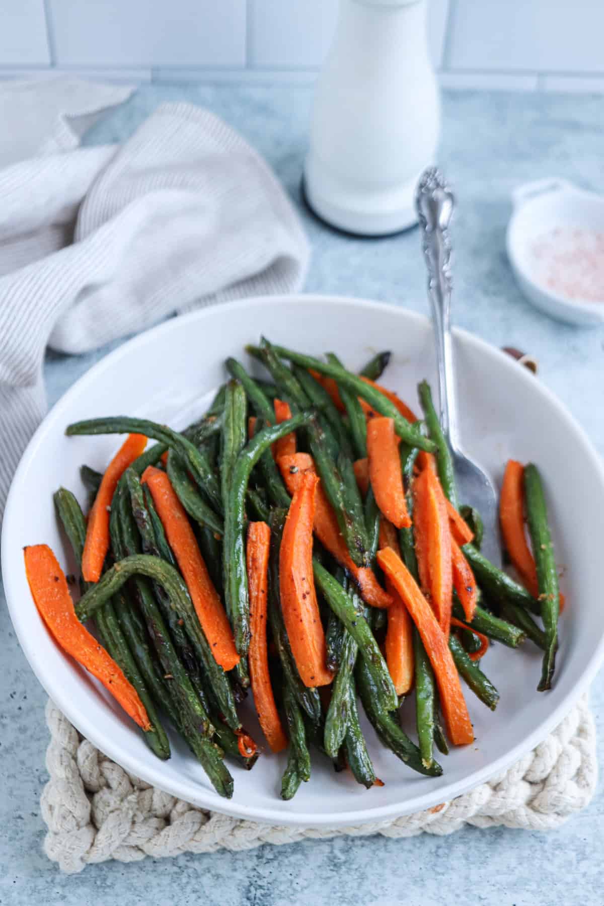 A white bowl filled with roasted green beans and carrot sticks sits on a blue surface, resembling a vibrant falafel platter. A spoon rests inside the bowl, accompanied by a striped cloth, a white pepper grinder, and a small dish with pink salt in the background.