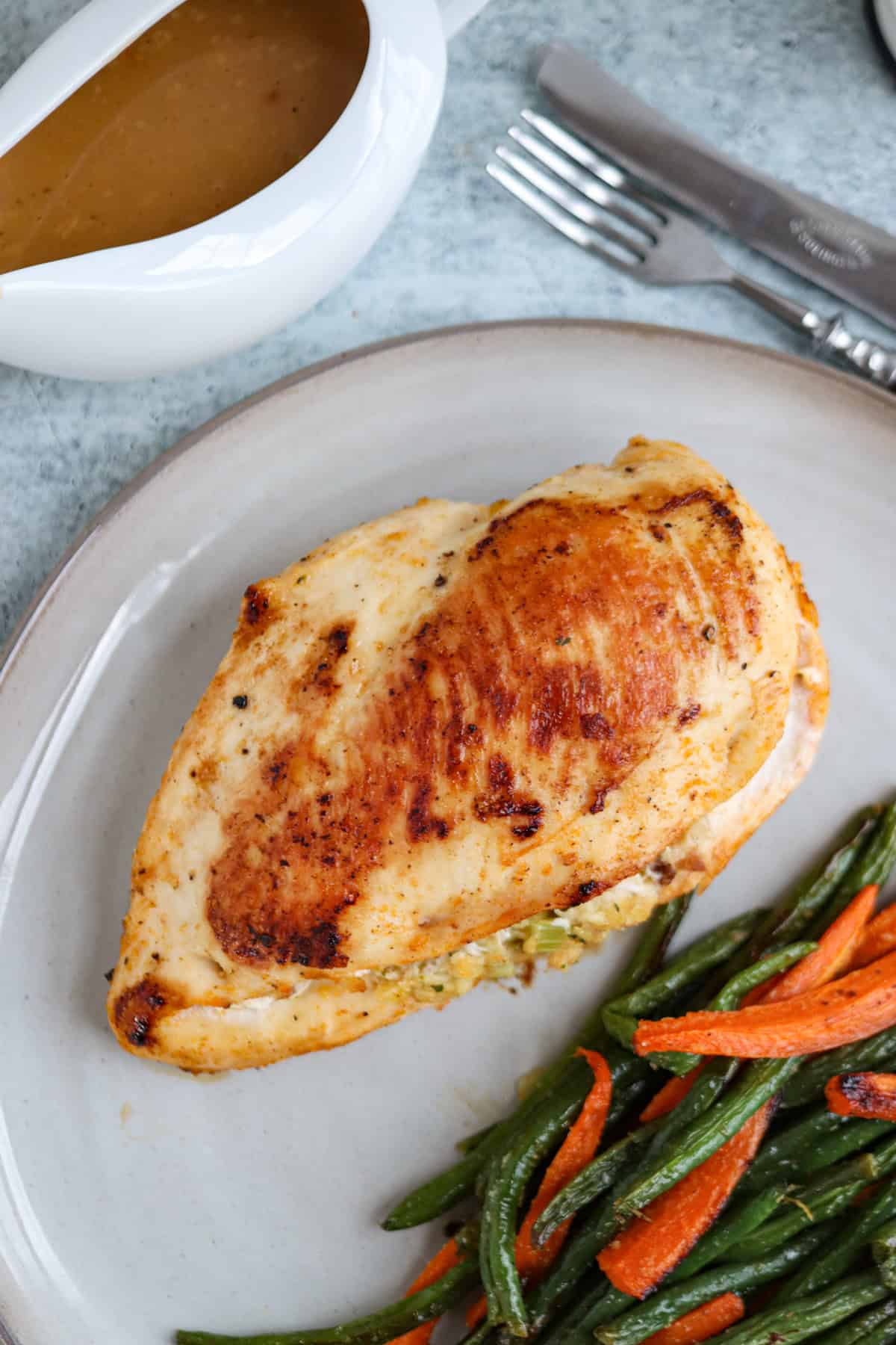 A cooked chicken breast on a plate alongside a vibrant medley of green beans and carrot slices. Nearby, a sauce boat brims with savory gravy, complemented by a fork and knife resting on the gray surface.