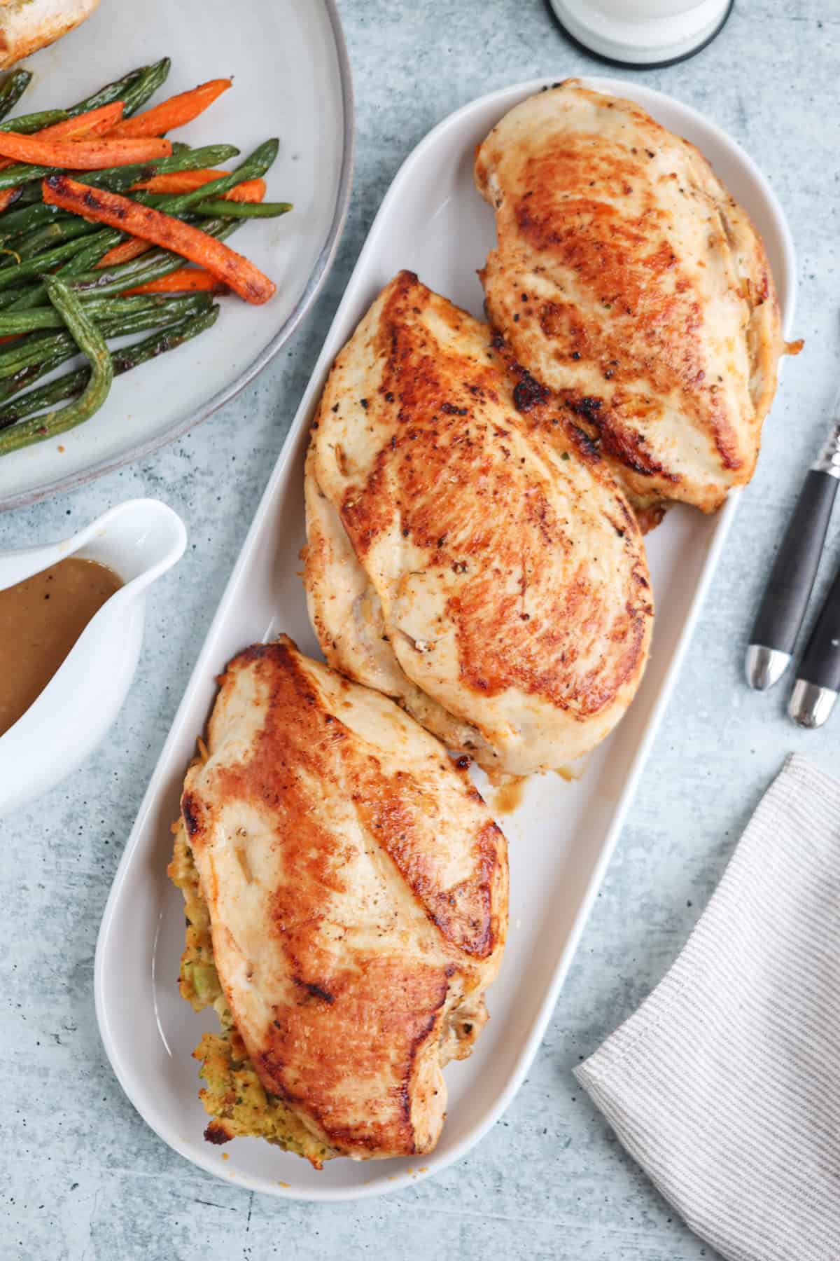 Three baked chicken breasts are served on a white platter. A side dish of roasted green beans and carrots is seen in the background, along with a gravy boat. A fork and knife rest on a napkin to the right.