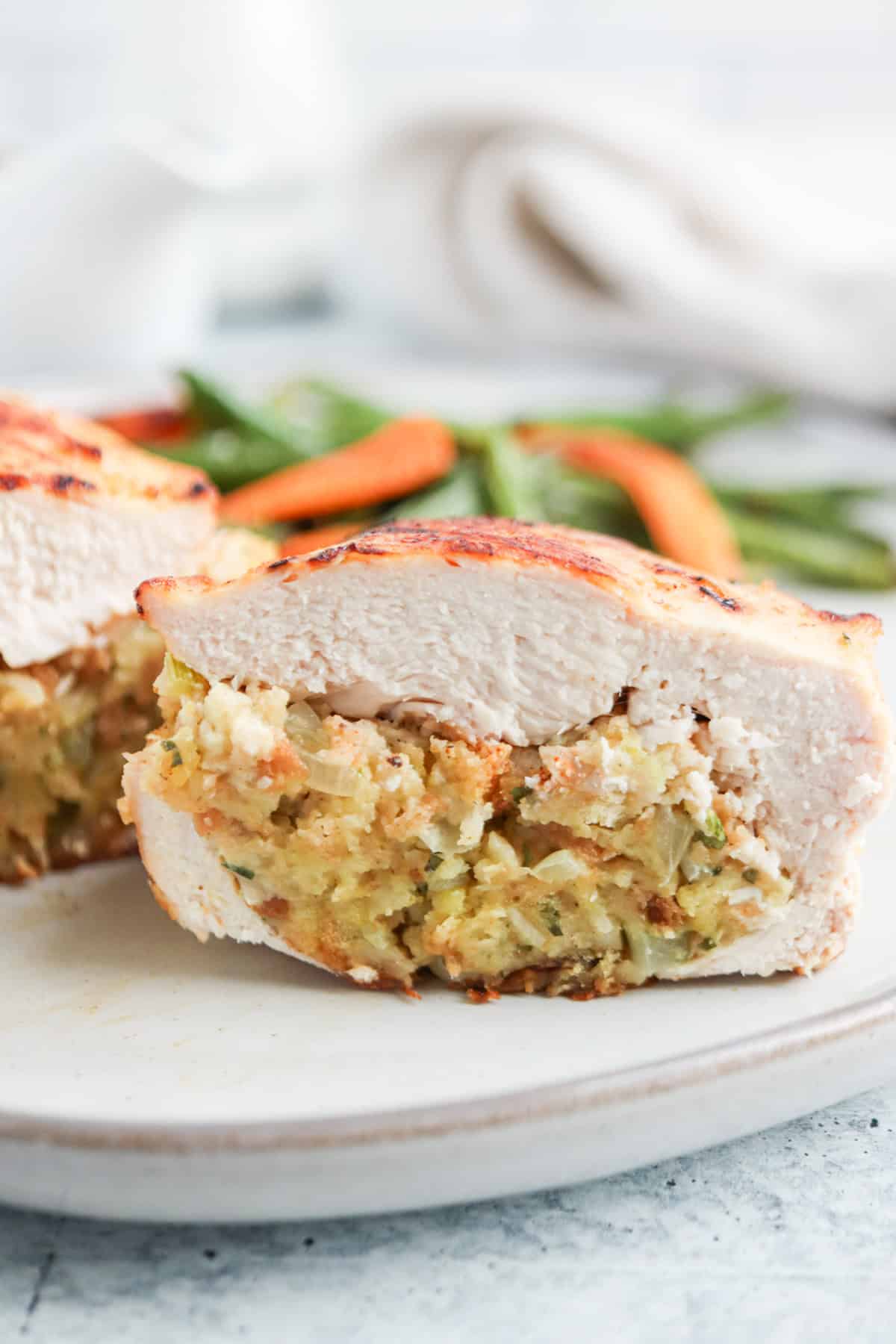 Close-up of a sliced stuffed chicken breast on a white plate, reminiscent of a falafel platter, filled with a herb and breadcrumb mixture. In the background, there are blurred cooked carrots and green beans.