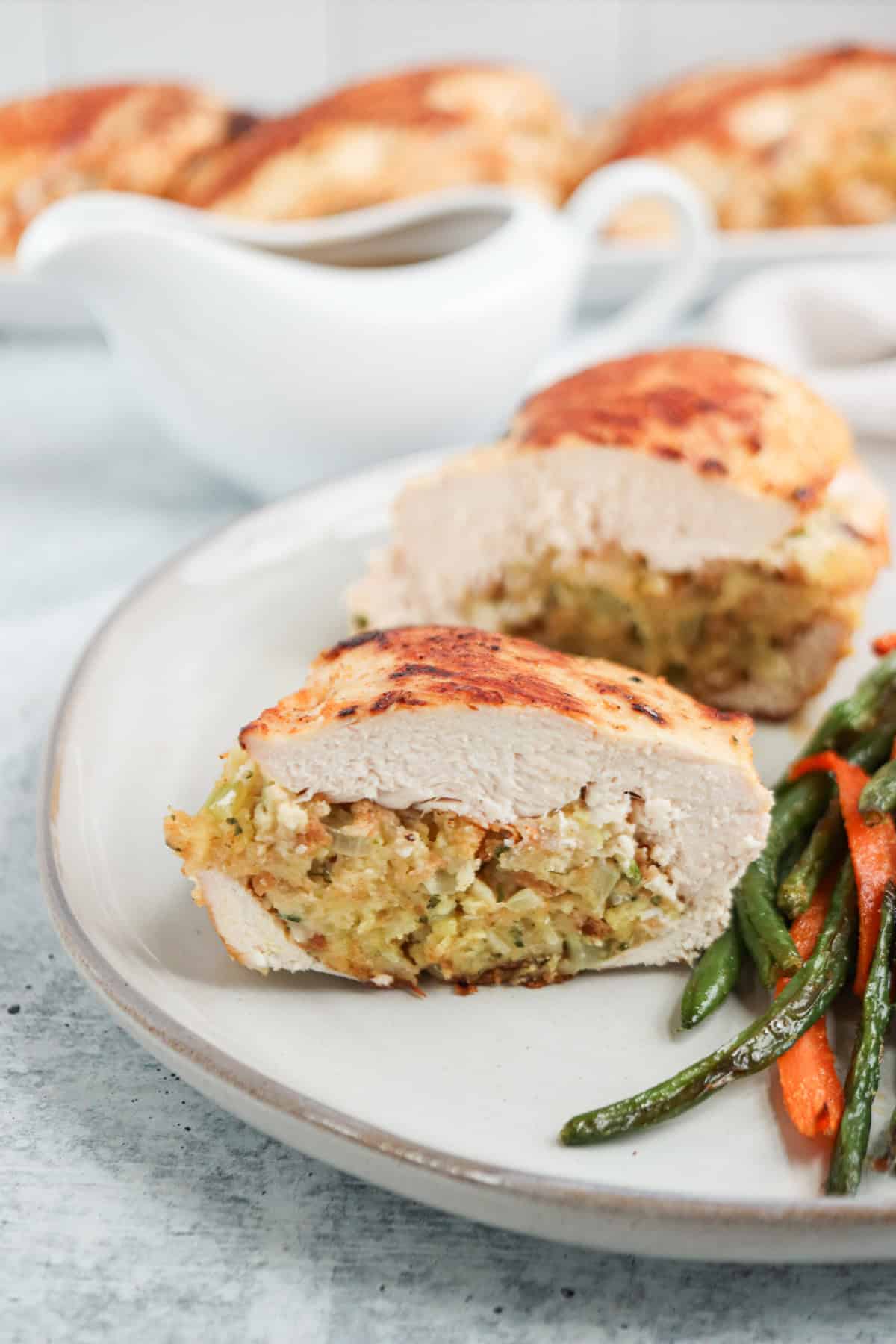 A plate featuring stuffed chicken breasts sliced open to reveal a flavorful filling. Sautéed green beans and carrots accompany this delicious duo, while a gravy boat in the background hints at an invitingly savory meal setting.