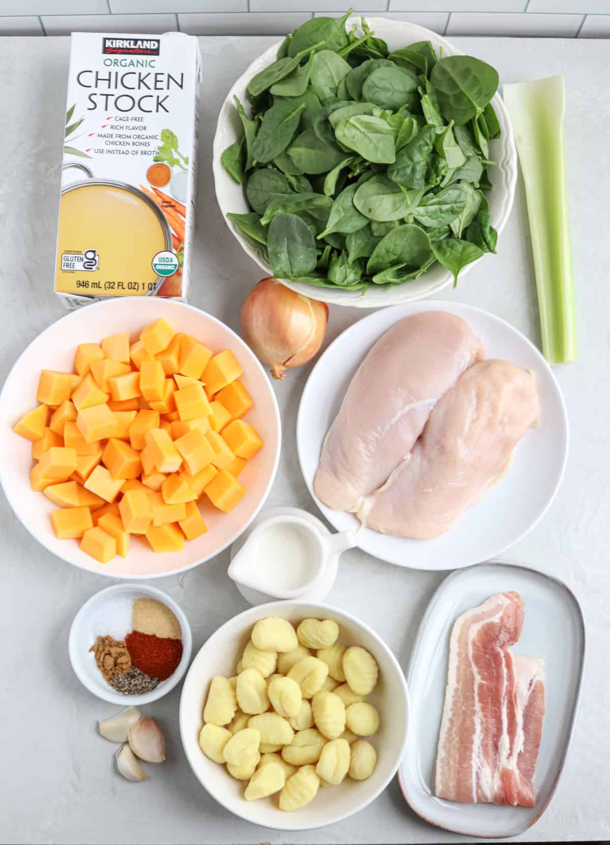 Ingredients for a chicken butternut squash soup are arranged on a surface, including organic chicken stock, fresh spinach, celery, onion, raw chicken breasts, diced butternut squash, gnocchi, spices, cream, and strips of bacon.