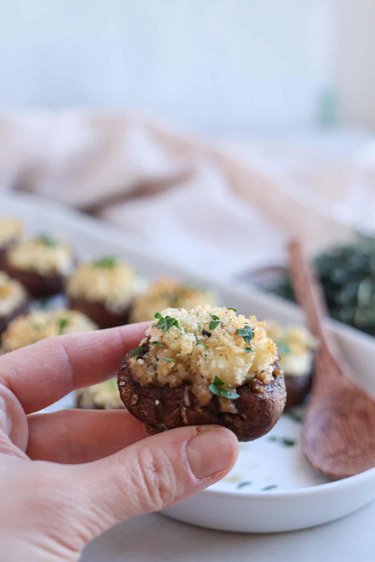 A hand holds a Boursin stuffed mushroom, topped with breadcrumbs and herbs. In the background, a platter brims with more of these delicious bites, alongside a wooden spoon and a striped cloth.