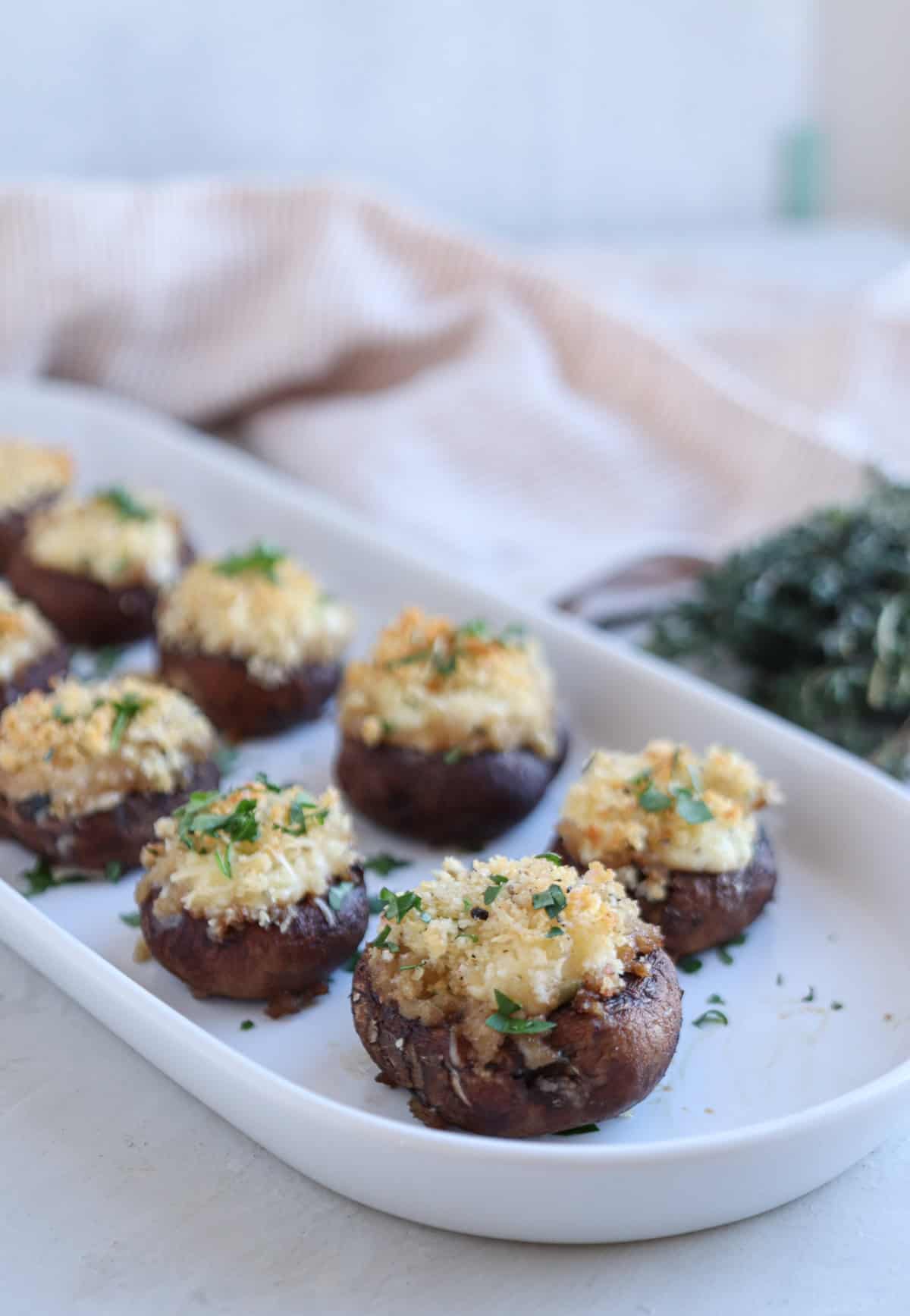 A white tray filled with Boursin stuffed mushrooms topped with breadcrumbs and herbs. A bunch of fresh herbs is blurred in the background, and a striped cloth is partially visible on the side. The setting is on a light surface.