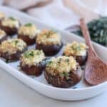 A white rectangular dish holds Boursin stuffed mushrooms topped with breadcrumbs and herbs. A wooden spoon lies beside them. The background features a cloth and a small bundle of herbs, adding to the rustic presentation.