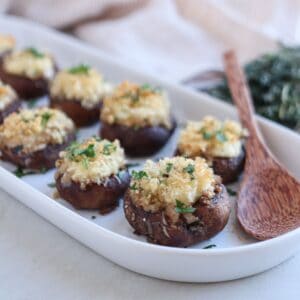 A white rectangular dish holds Boursin stuffed mushrooms topped with breadcrumbs and herbs. A wooden spoon lies beside them. The background features a cloth and a small bundle of herbs, adding to the rustic presentation.