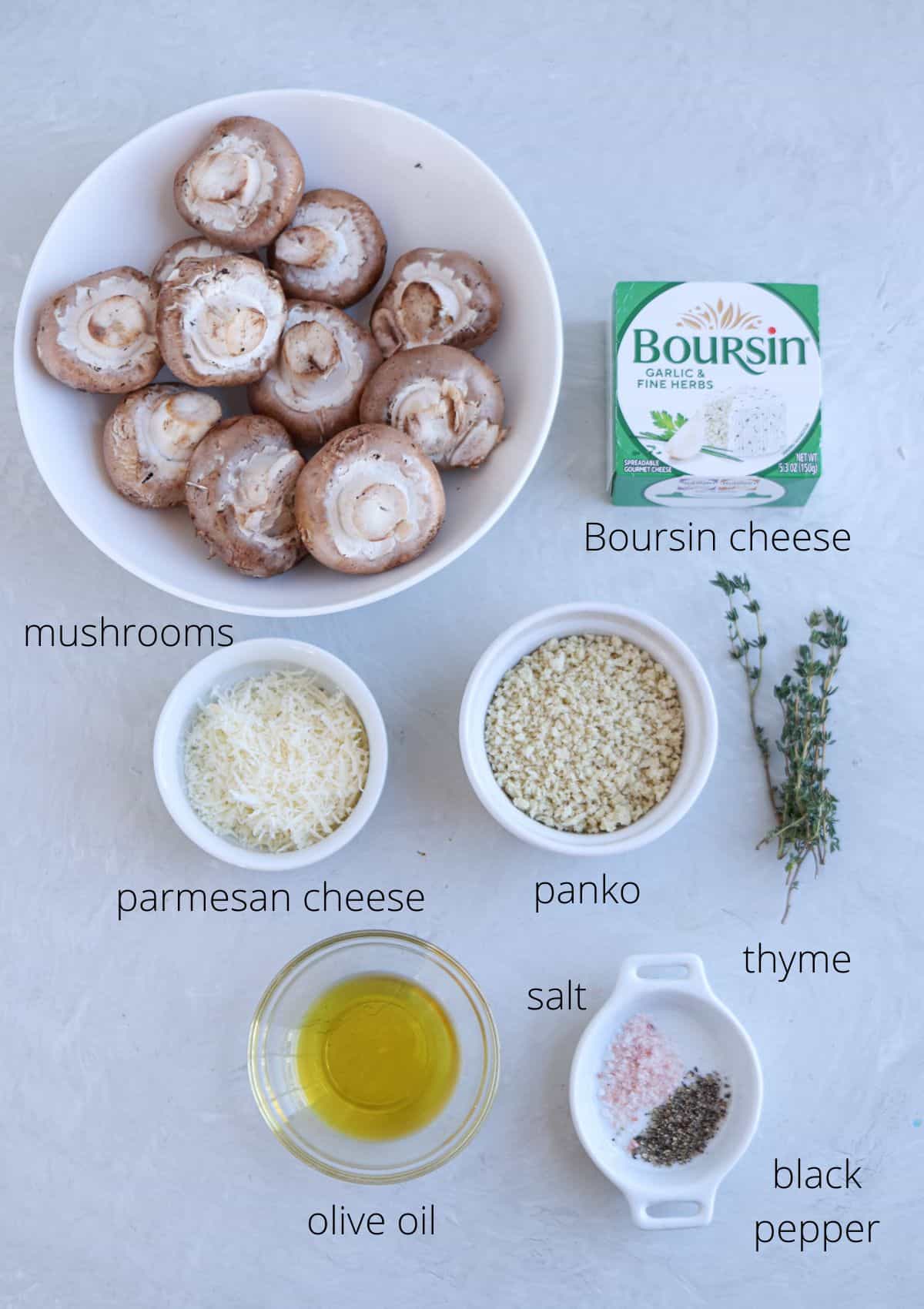 Overhead view of ingredients on a light surface, featuring a bowl of mushrooms, perfect for Boursin stuffed mushrooms. Surrounding are small bowls of parmesan cheese, panko, olive oil, salt, and pepper, with sprigs of thyme for that extra touch.