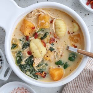 A creamy chicken butternut squash soup filled with gnocchi, shredded chicken, and vibrant chunks of orange squash. Wilted greens and bits of red pepper complement it. A spoon rests on the bowl's edge, with pink salt and pepper in small dishes beside it for seasoning perfection.