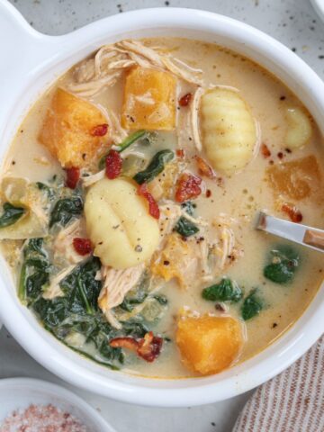 A creamy chicken butternut squash soup filled with gnocchi, shredded chicken, and vibrant chunks of orange squash. Wilted greens and bits of red pepper complement it. A spoon rests on the bowl's edge, with pink salt and pepper in small dishes beside it for seasoning perfection.