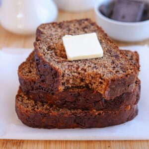 Three slices of banana bread stacked on a white plate with a pat of butter on top, resembling a delightful falafel platter. The bread is moist with visible banana and chocolate flecks. A white bowl with chocolate pieces graces the background on a wooden surface.