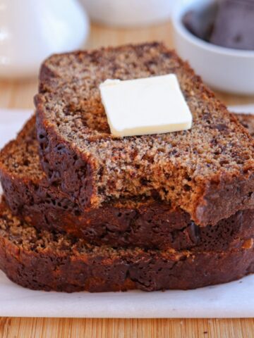 Three slices of banana bread stacked on a white plate with a pat of butter on top, resembling a delightful falafel platter. The bread is moist with visible banana and chocolate flecks. A white bowl with chocolate pieces graces the background on a wooden surface.