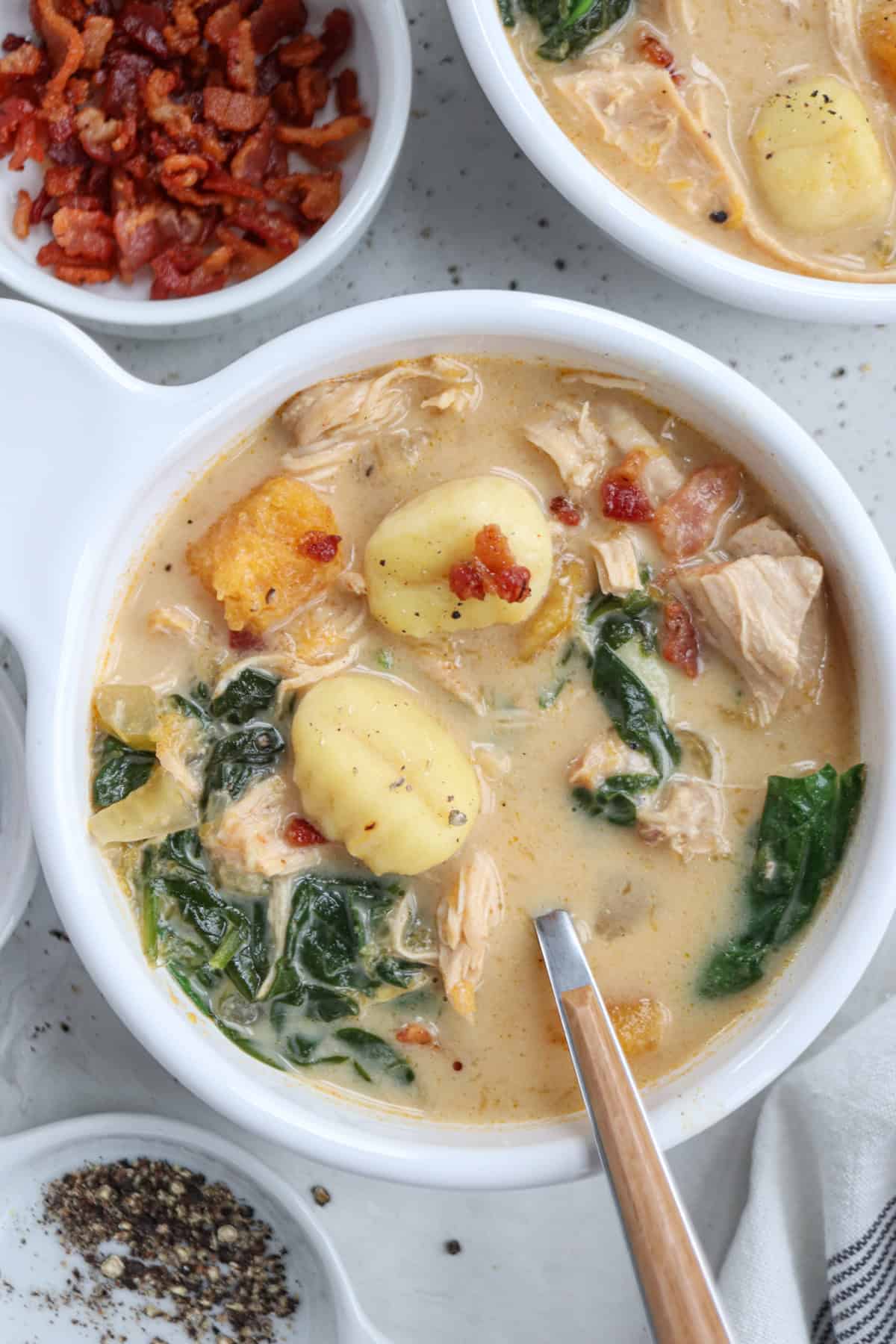 A bowl of creamy chicken butternut squash soup with gnocchi, shredded chicken, spinach, and crispy bacon bits. Another bowl is partially visible, with a side dish of bacon and pepper nearby. A spoon rests in the bowl.