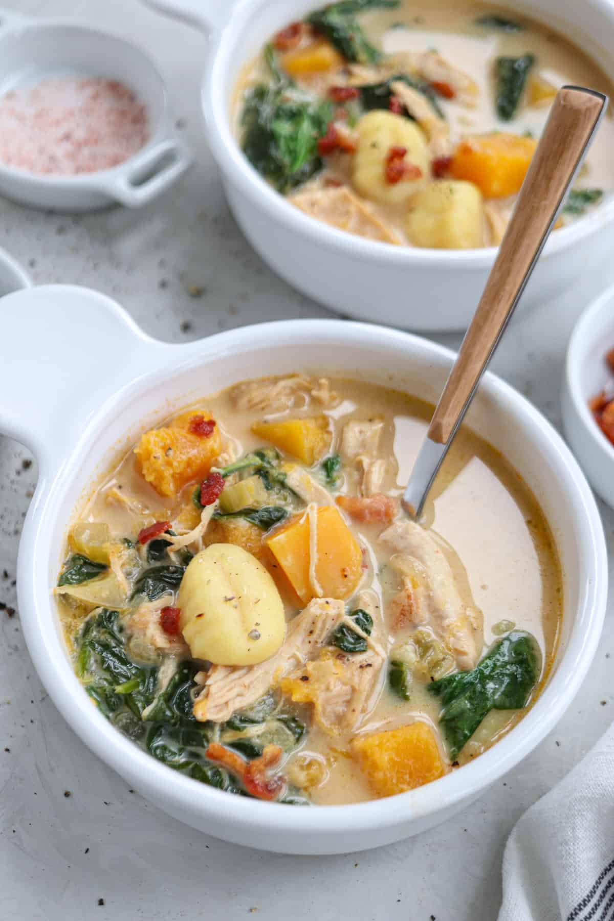A creamy chicken butternut squash soup in a white bowl brims with tender chicken, potatoes, and vibrant spinach. A wooden-handled spoon rests inside, ready to be savored. In the background, another bowl of this delightful soup awaits. Nearby, small dishes of pink salt and spices add a finishing touch.
