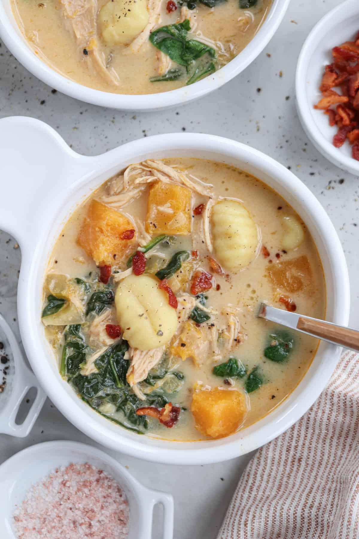 A bowl of creamy chicken and butternut squash soup with gnocchi, spinach, and chunks of butternut squash. The soup is garnished with pepper and served with a side of crispy bacon bits. Seasonings are scattered around a striped cloth nearby.