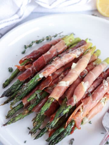 A plate of prosciutto-wrapped asparagus, garnished with fresh thyme, rests elegantly on a white platter. Accompanied by a lemon wedge and a white napkin in the background, this dish is an inviting blend of flavors and presentation.