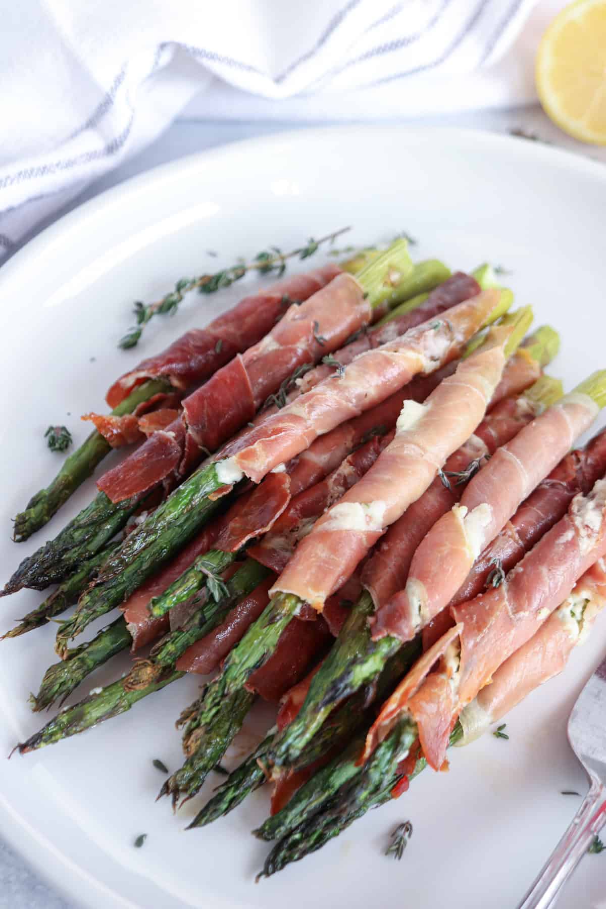 A plate of prosciutto-wrapped asparagus, garnished with fresh thyme, rests elegantly on a white platter. Accompanied by a lemon wedge and a white napkin in the background, this dish is an inviting blend of flavors and presentation.