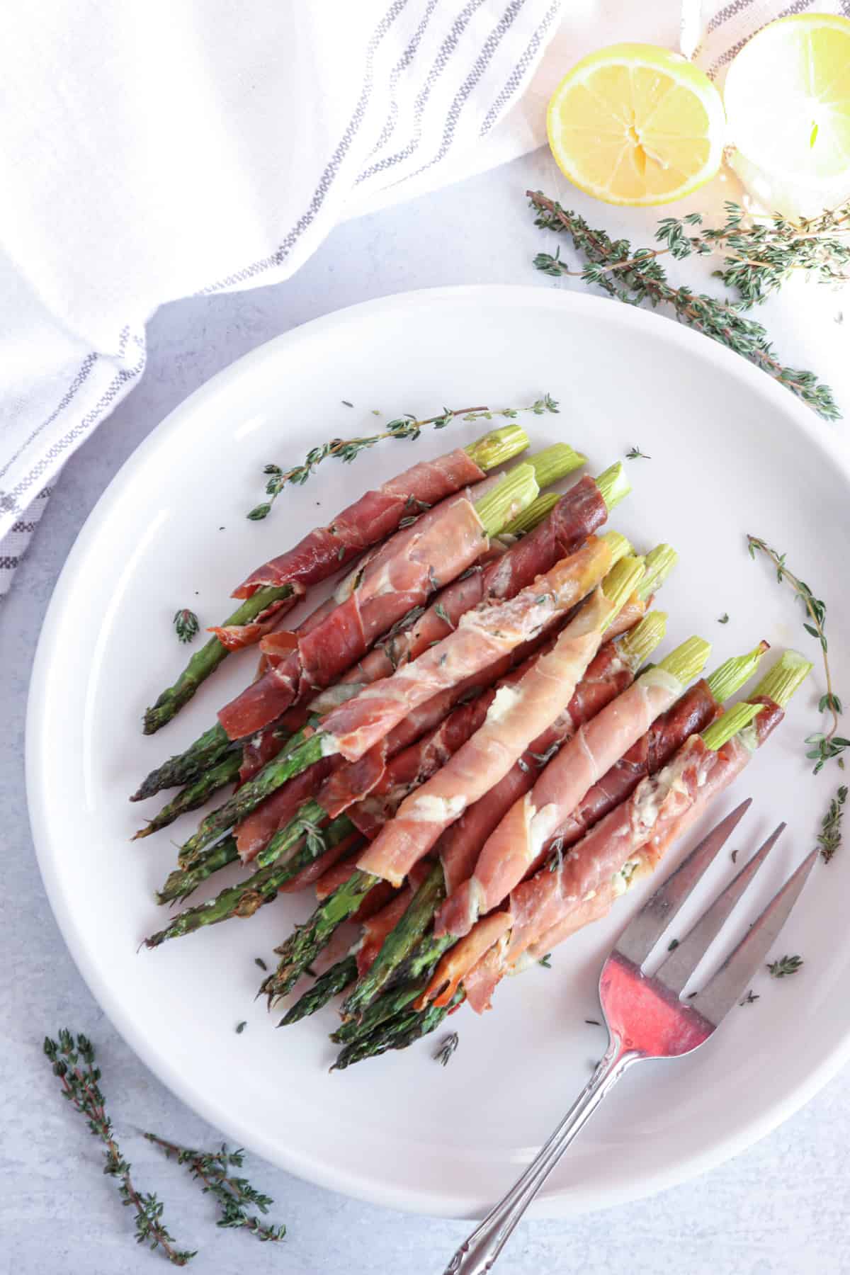 A plate of prosciutto-wrapped asparagus garnished with fresh thyme, with a fork on the side. The background features a white napkin and lemon slices.