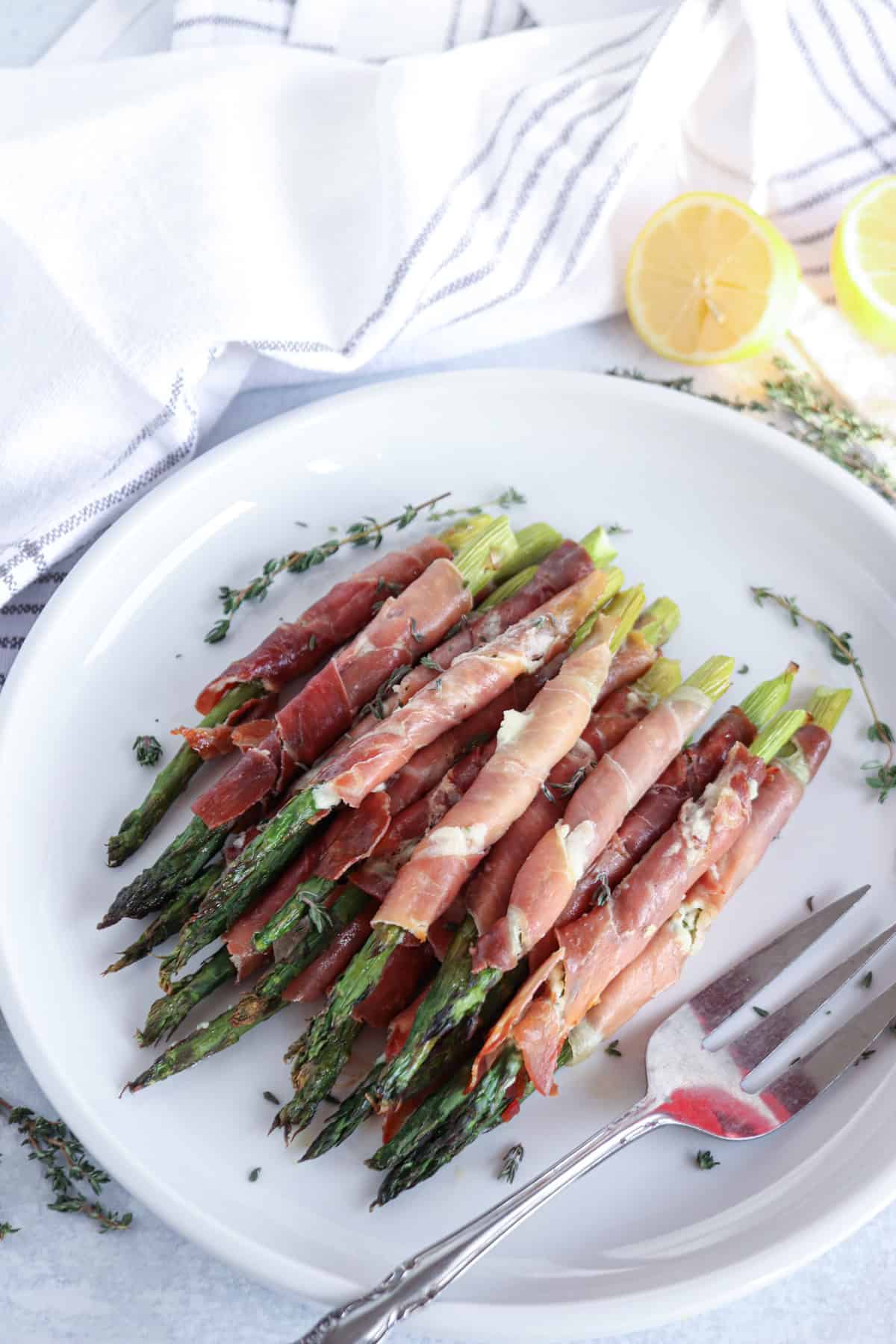 A white plate showcases prosciutto wrapped asparagus, elegantly garnished with herbs. A fork rests invitingly on the plate. In the background, vibrant sliced lemons complement a white and gray striped cloth.