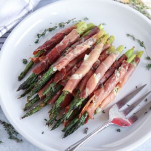 A plate of prosciutto-wrapped asparagus, garnished with herbs. A fork rests beside the neatly arranged bundles, ready to savor this delightful arrangement.