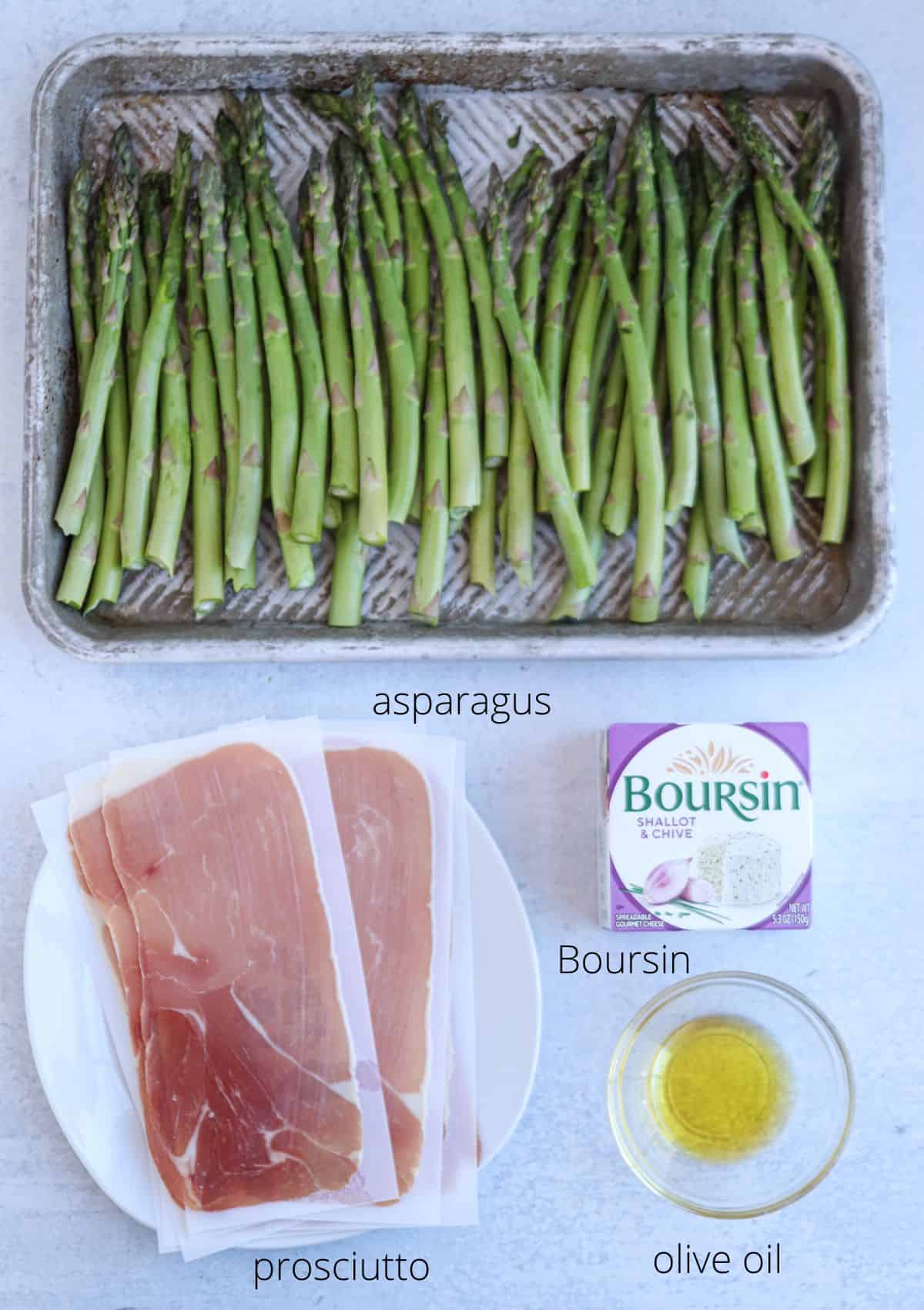 A cooking preparation scene features prosciutto-wrapped asparagus meticulously arranged on a baking tray, accompanied by a plate with more prosciutto slices, a small bowl of olive oil, and a package of Boursin cheese on a light-colored surface.