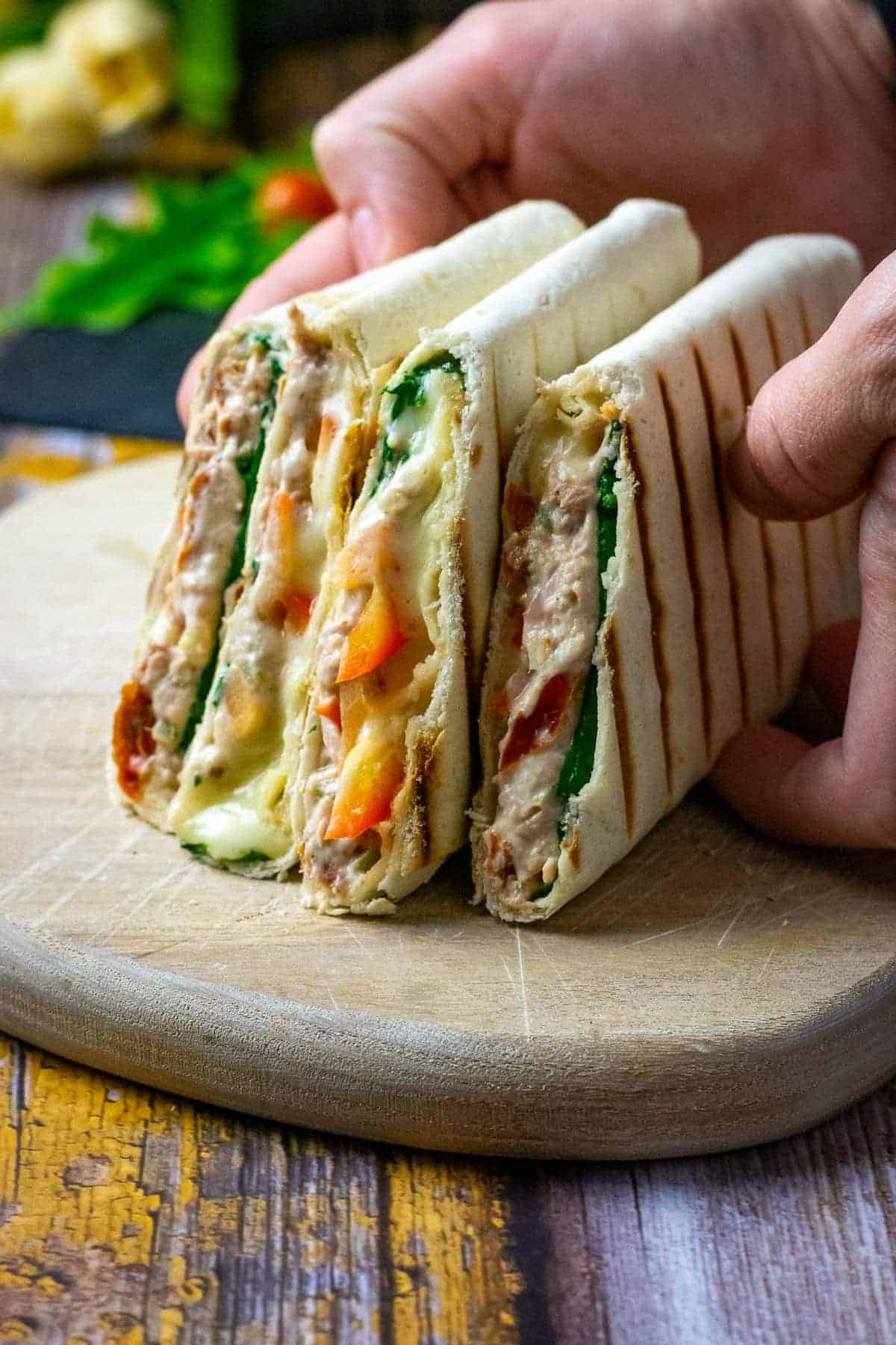 Three folded tortillas filled with creamy chicken, spinach, tomatoes, and melted cheese are being held on a wooden board. The tortillas have grill marks, and the background includes fresh vegetables.