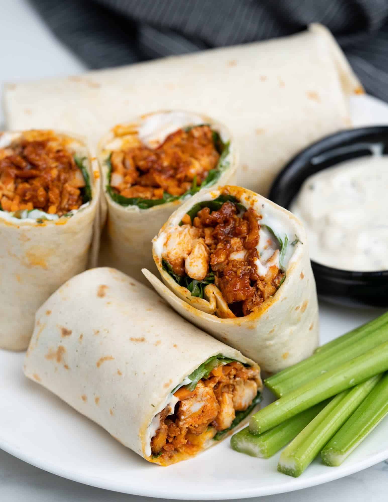 Four chicken wraps filled with shredded chicken, greens, and sauce are arranged on a plate. A small bowl of dipping sauce and celery sticks accompany the wraps. A striped cloth is partially visible in the background.