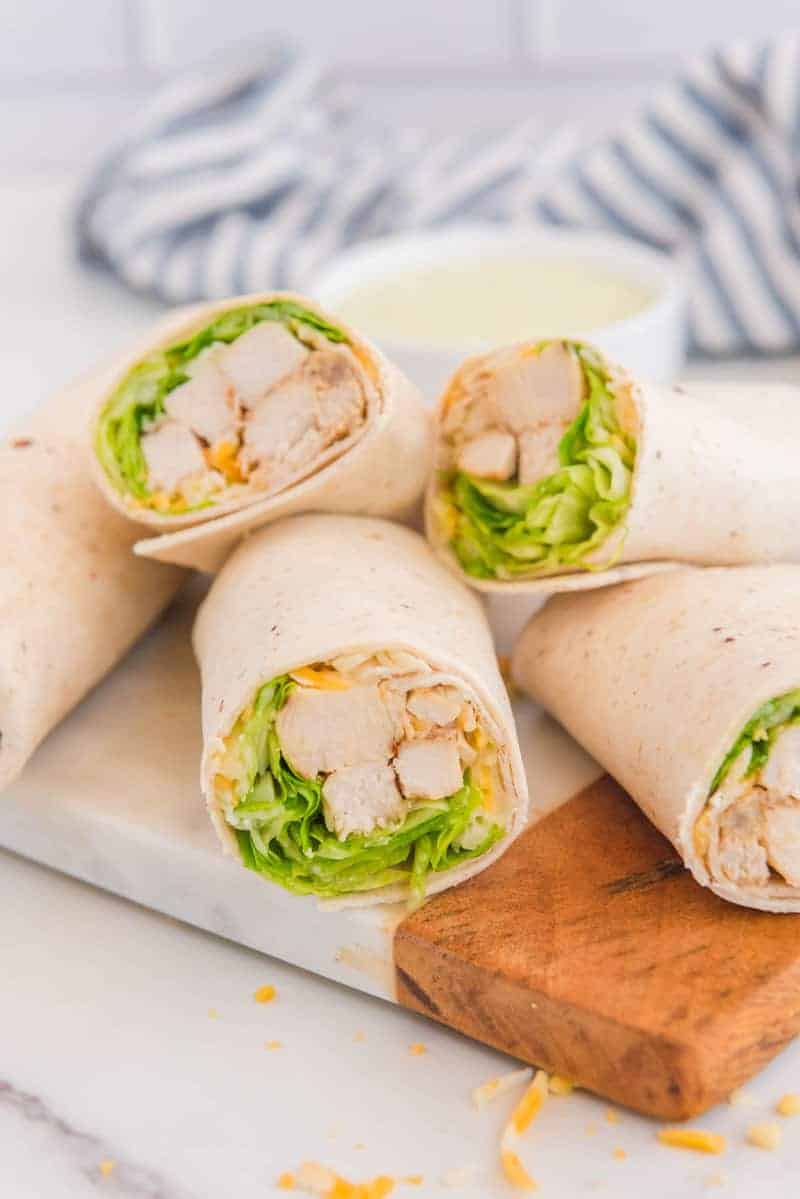Chicken wraps filled with lettuce, cheese, and diced chicken are stacked on a marble board. A striped napkin and a bowl of sauce are blurred in the background.