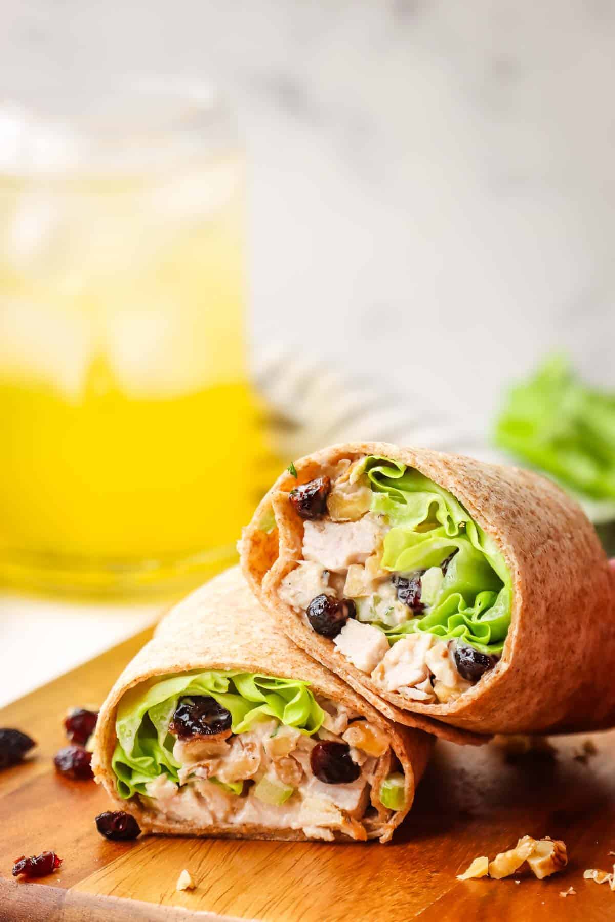 Two halves of a wrap filled with lettuce, chicken, walnuts, and cranberries sit on a wooden board. A blurred glass of a yellow drink is in the background.