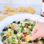 A hand dips a tortilla chip into a vibrant taco dip, enhanced with cottage cheese, shredded cheese, sliced olives, diced tomatoes, chopped avocados, and herbs. In the background, scattered tortilla chips accompany the tantalizing sight of a hot sauce bottle.