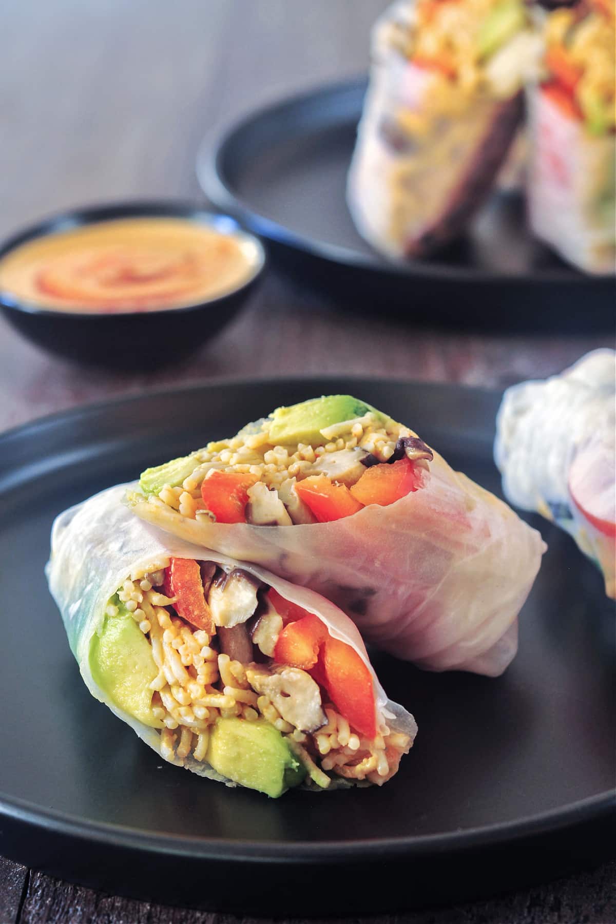 Close-up of fresh spring rolls filled with colorful vegetables, nuts, and noodles, sliced and displayed on a dark plate. A small dish of dipping sauce is in the background on a wooden surface.