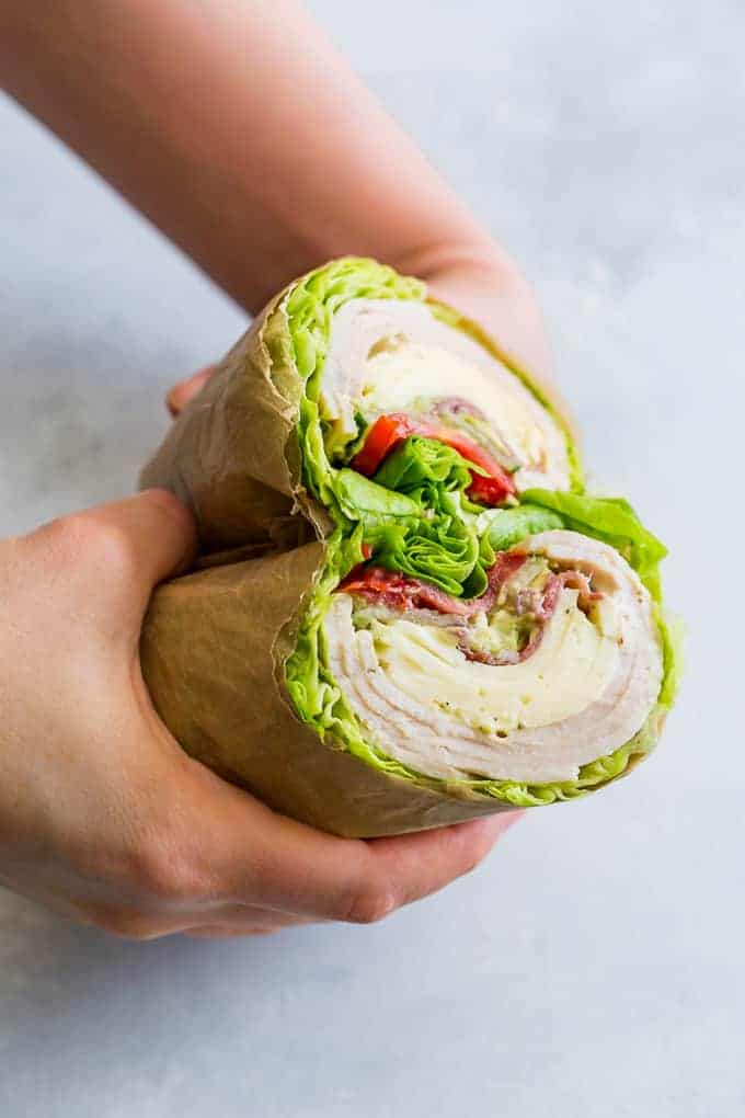 Hands holding a wrap sandwich cut in half, wrapped in brown paper. The sandwich contains turkey, lettuce, tomato, and cheese, with some fillings visible in cross-section. The background is a light gray surface.