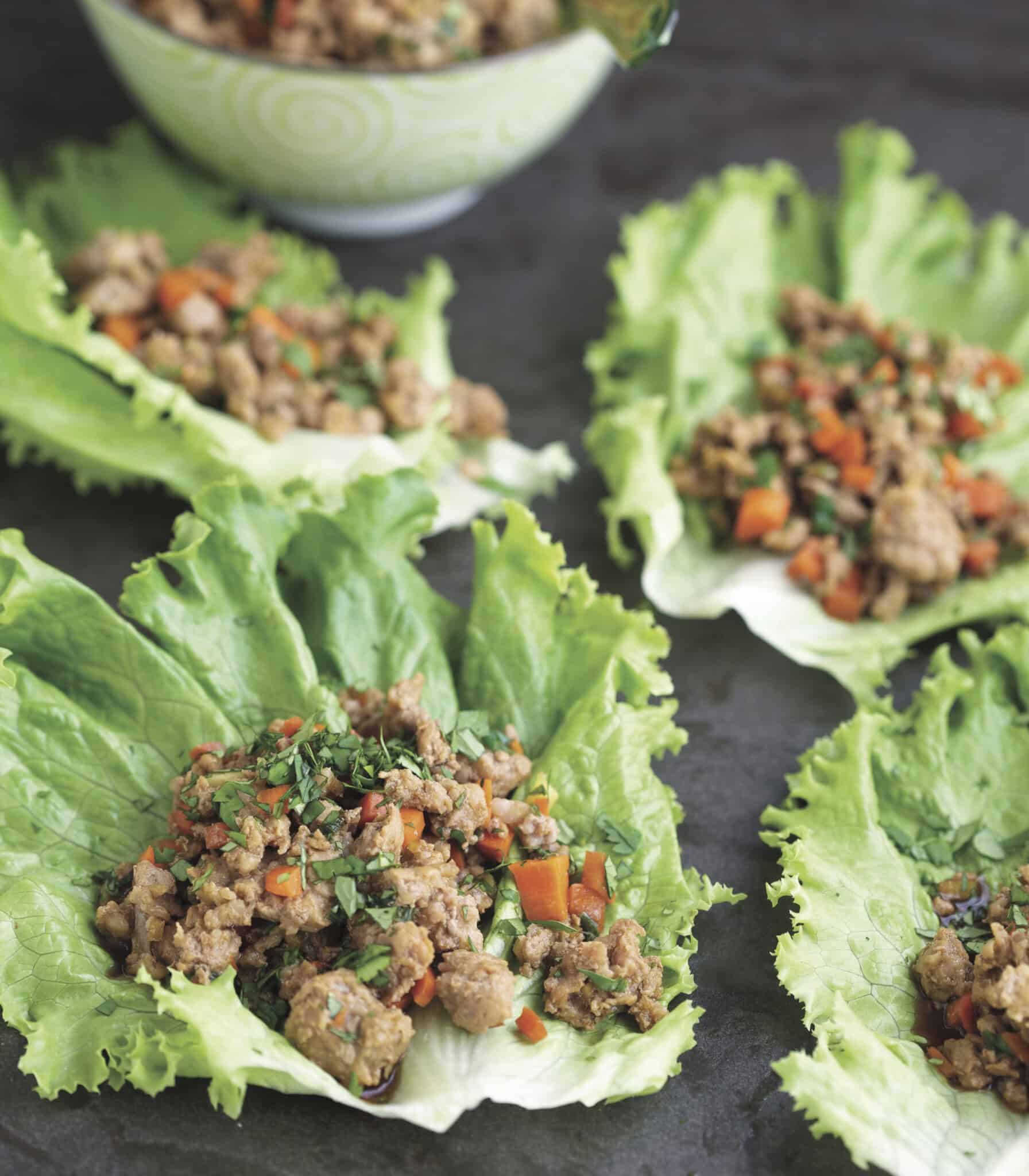 Lettuce wraps filled with cooked ground meat, diced carrots, and herbs are arranged on a dark surface. A decorative bowl in the background is partially visible.
