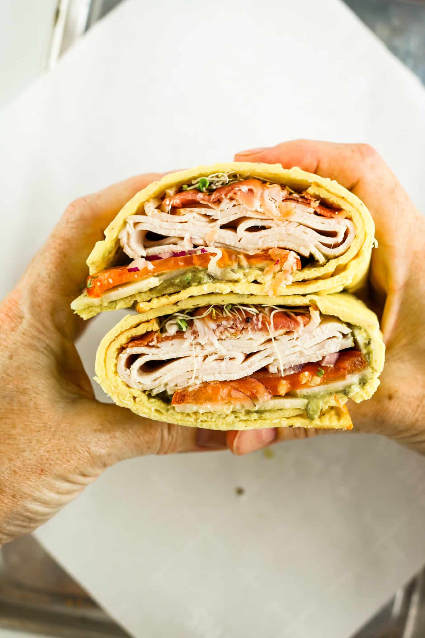 Two hands holding a halved wrap sandwich filled with slices of turkey, tomato, greens, and sauce. The sandwich is layered neatly, showing the colorful ingredients inside. It is set against a plain background.