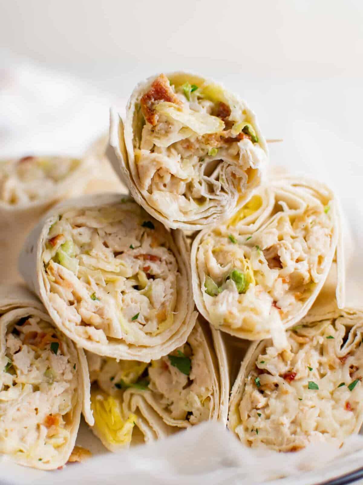 A close-up of several chicken salad wraps cut in halves, showcasing a filling of shredded chicken, lettuce, and herbs, neatly arranged on a white plate. The wraps have a light, fresh appearance with visible layers of ingredients.