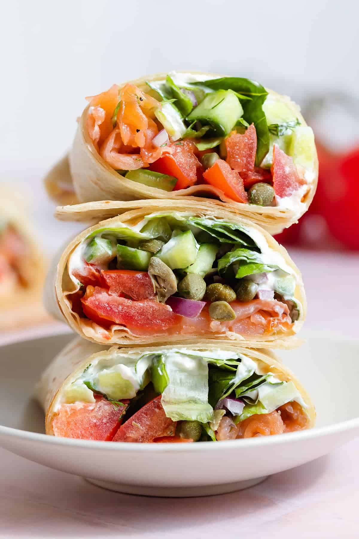 Three stacked wraps filled with smoked salmon, sliced cucumbers, tomatoes, capers, red onions, and lettuce, served on a white plate. The wraps are cut in half, showcasing the colorful and fresh ingredients inside.