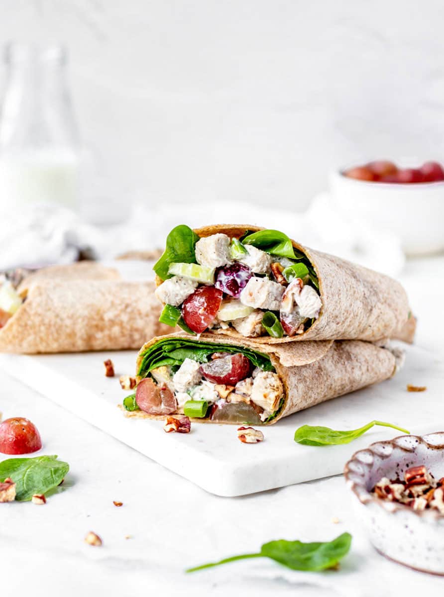 A close-up of a wrap filled with chicken, grapes, spinach, and nuts, sliced in half and placed on a white board. Surrounding the wrap are scattered spinach leaves and chopped nuts, with a small bowl of grapes blurred in the background.