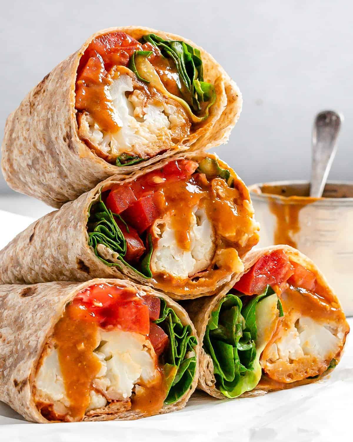 Four stacked wraps filled with crispy cauliflower, fresh spinach, and diced tomatoes, drizzled with a creamy brown sauce. A small bowl with more sauce and a spoon sits in the background.