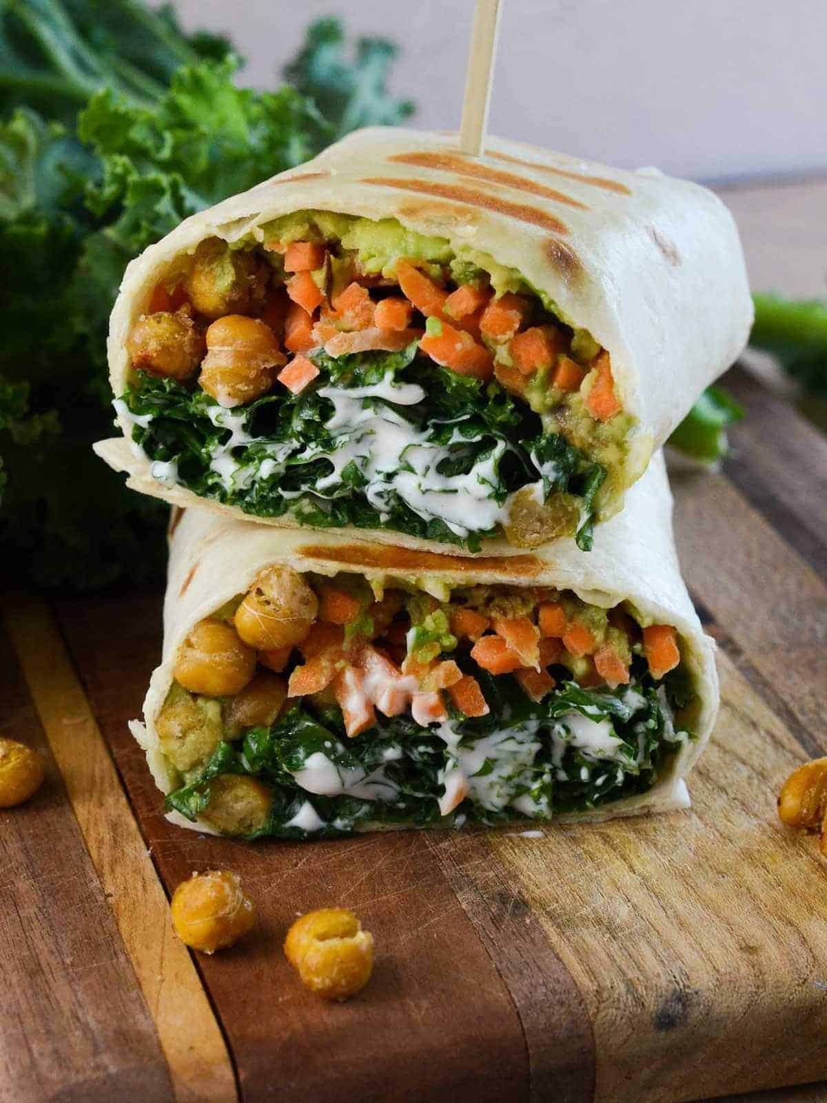 A wrap cut in half and stacked, filled with roasted chickpeas, shredded carrots, kale, creamy dressing, and guacamole, on a wooden cutting board. Extra chickpeas and kale are scattered around.