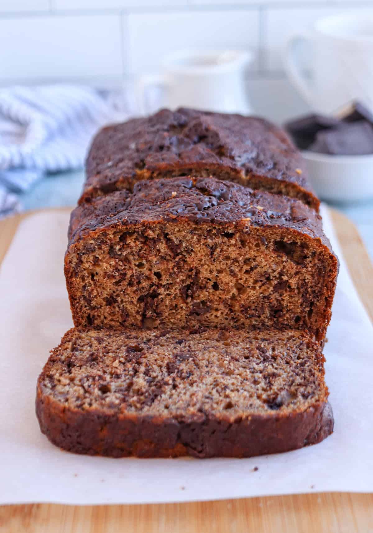 A sliced loaf of banana bread with a golden-brown crust rests on a wooden cutting board, its texture speckled with dark bits of chocolate chips. 
