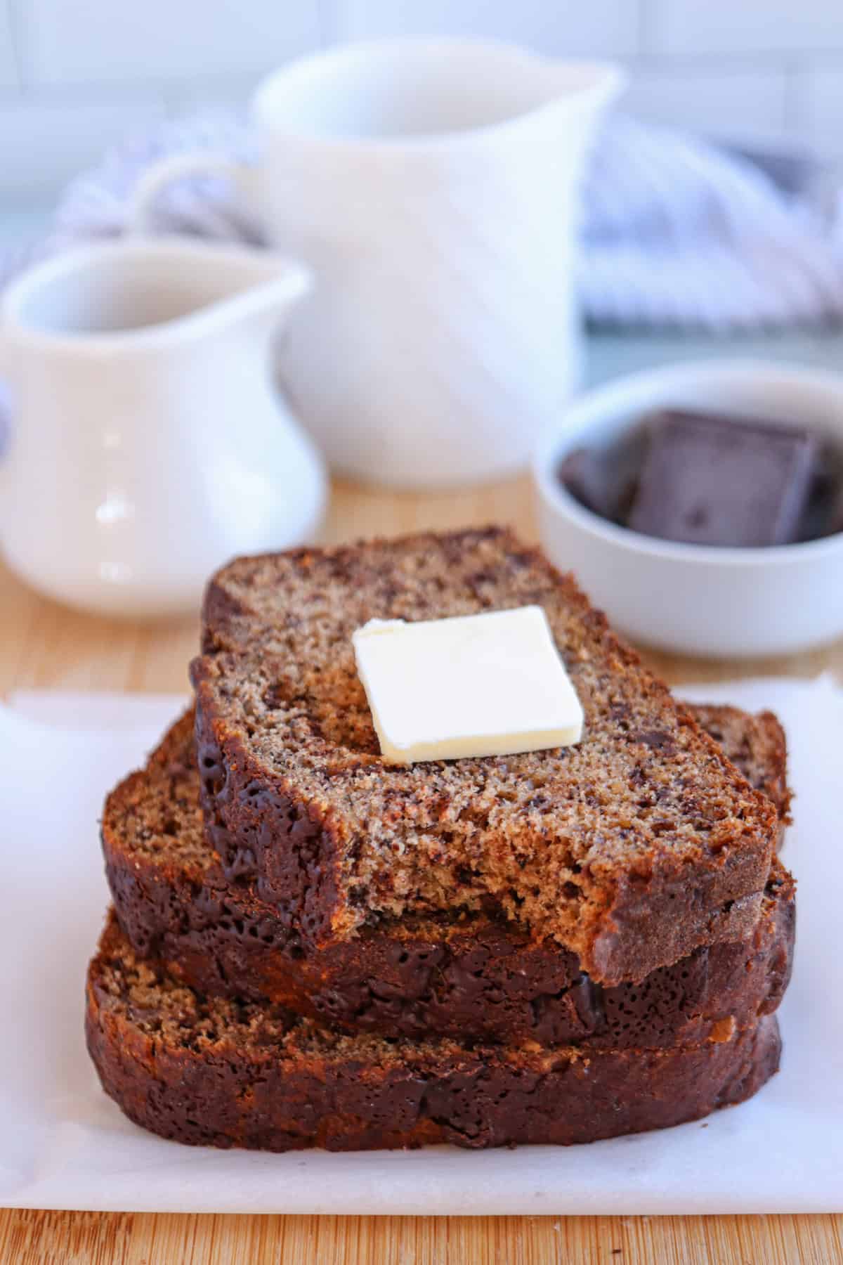 Three slices of banana bread stacked on white parchment paper, topped with a pat of butter. In the background, alongside a white jug and a striped cloth, is a bowl with chocolate pieces.
