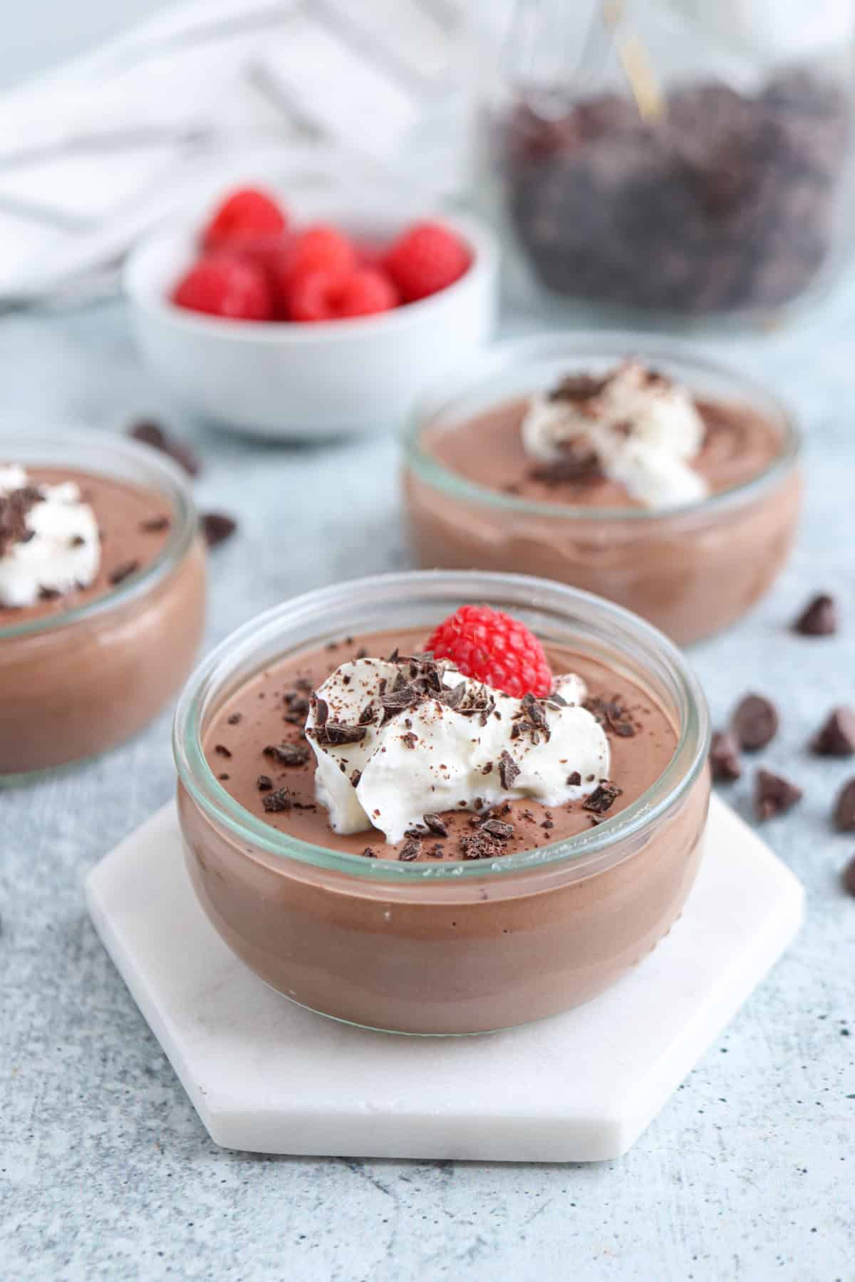 Three ramekins of chocolate mousse topped with whipped cream, chocolate shavings, and a raspberry are arranged on a hexagonal marble coaster. In the background, there is a bowl of fresh raspberries and a jar of chocolate chips.