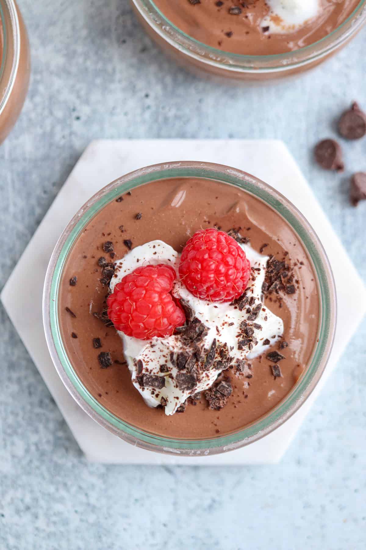A glass bowl of chocolate mousse topped with whipped cream, two raspberries, and chocolate shavings. The bowl is on a hexagonal white coaster, with chocolate chips scattered around on a gray surface.