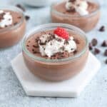A small glass jar of chocolate mousse topped with whipped cream, chocolate shavings, and a raspberry. The jar is placed on a hexagonal white coaster. More jars and chocolate chips are blurred in the background on a light gray surface.
