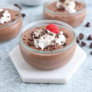 A small glass jar of chocolate mousse topped with whipped cream, chocolate shavings, and a raspberry. The jar is placed on a hexagonal white coaster. More jars and chocolate chips are blurred in the background on a light gray surface.