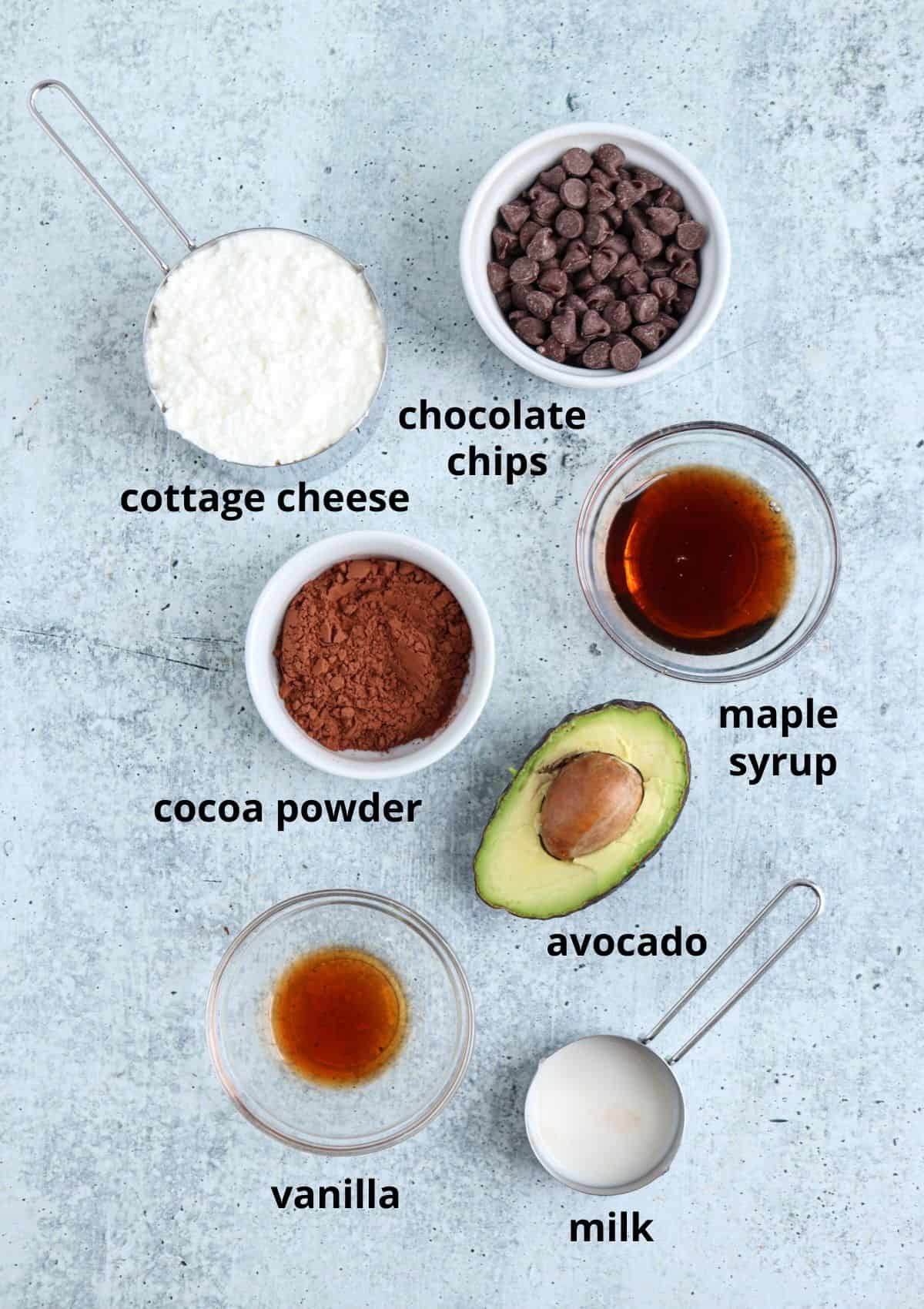 A flat lay of seven ingredients labeled on a textured surface: cottage cheese, chocolate chips, cocoa powder, maple syrup, avocado, vanilla, and milk. Each is in separate bowls or containers.