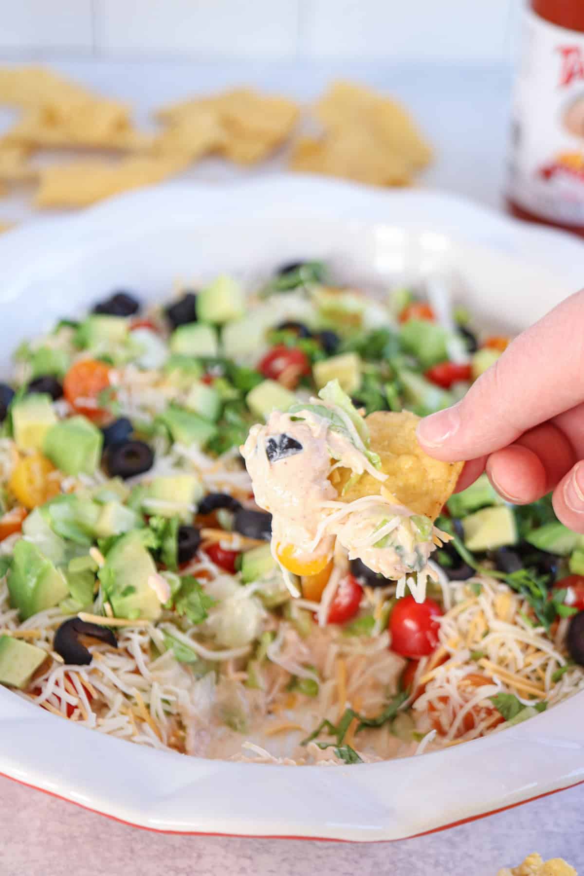 A hand holds a tortilla chip scooping a layered taco dip topped with diced avocado, cherry tomatoes, sliced olives, and shredded cheese. Cottage cheese adds a creamy twist to this savory blend. Chips and a bottle of hot sauce linger in the background.